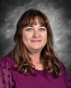 A woman with long, brown hair and a purple blouse smiles in front of a gray background.