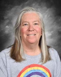 Smiling woman with long hair wearing a gray shirt with a rainbow design, set against a cloudy background.