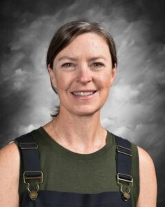 A smiling woman with short brown hair, wearing a green shirt and dark overalls, against a gray background.