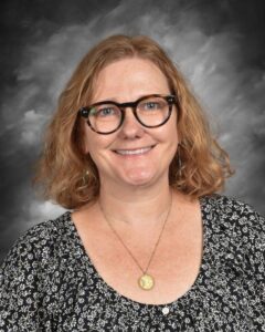 A woman with curly hair and glasses smiles, wearing a floral top and a necklace, against a gray backdrop.