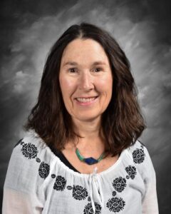 A woman with long, wavy hair smiles, wearing a white embroidered blouse and a colorful necklace, against a gray backdrop.
