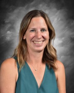 Smiling woman with shoulder-length brown hair, wearing a teal top, against a gray background.