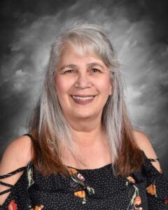 Smiling woman with long gray hair wearing a floral black top, set against a gray background.