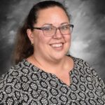 Smiling woman with glasses, wearing a patterned blouse, against a gray background.