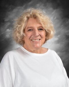 Smiling woman with curly blonde hair wearing a white shirt, against a gray background.
