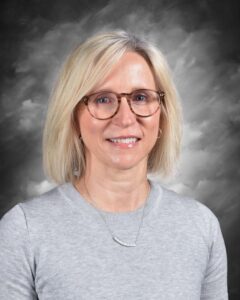 A smiling woman with short blonde hair and glasses, wearing a gray sweater, against a cloudy gray background.