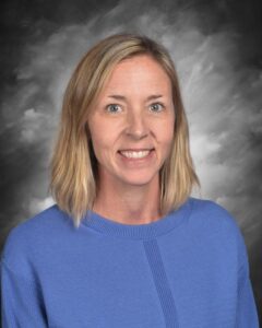 A smiling woman with shoulder-length blonde hair, wearing a blue sweater, set against a gray background.