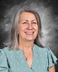 Smiling woman with shoulder-length gray hair, wearing a teal blouse, against a gray background.