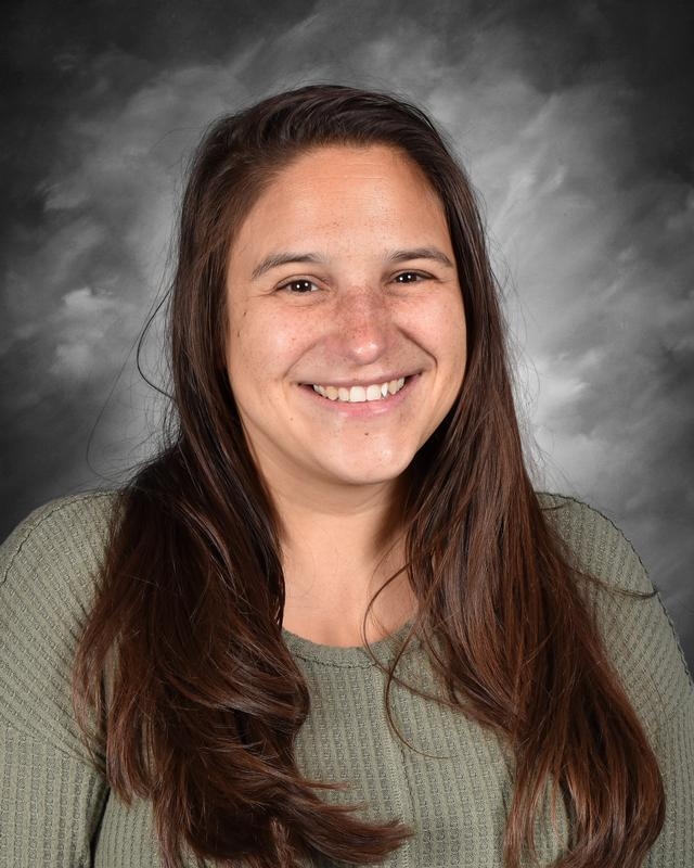 A smiling woman with long brown hair wearing a green sweater, set against a soft gray background.