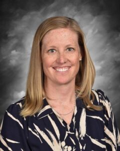 Smiling woman with shoulder-length blonde hair, wearing a patterned navy blouse, against a gray background.