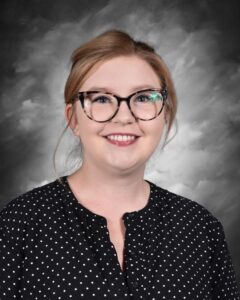 Smiling woman with glasses and blonde hair, wearing a black polka dot blouse, against a gray background.