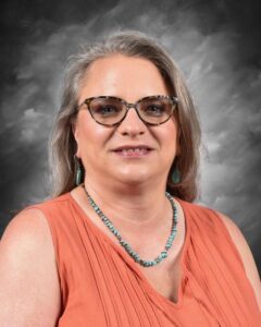 A woman with glasses and long hair smiles, wearing an orange top and a turquoise necklace against a gray background.