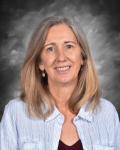 Smiling woman with long, straight hair wearing a light blue striped shirt and a maroon top, against a gray background.