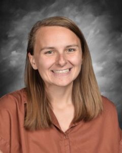 Smiling woman with long brown hair wearing a brown shirt, set against a gray background.