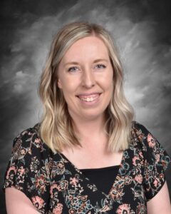 Smiling woman with wavy blonde hair, wearing a floral black top, against a gray background.