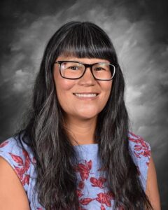 Smiling woman with long black hair and glasses, wearing a floral-patterned blue top, against a gray background.
