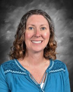 Smiling woman with curly hair wearing a blue shirt with embroidery, against a gray backdrop.