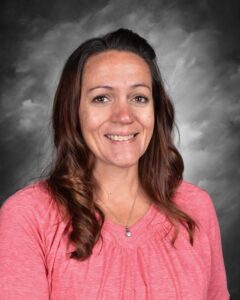 Smiling woman with long, wavy brown hair, wearing a pink shirt, against a gray background.