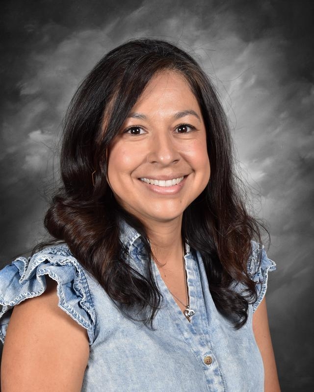 A smiling woman with long, dark hair wearing a denim shirt with ruffled sleeves, set against a gray background.