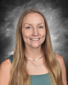 Headshot of a woman with long, straight blonde hair, wearing a teal top and a silver necklace, smiling against a dark background.