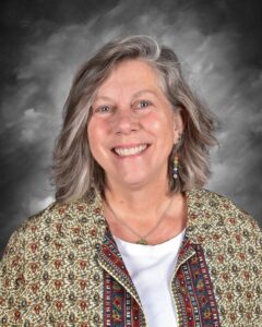 A smiling woman with shoulder-length gray hair, wearing a patterned jacket over a white shirt, against a gray backdrop.