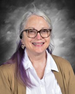 Smiling woman with glasses and purple hair, wearing a white shirt and tan cardigan, against a gray background.