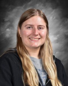 A smiling young woman with long, straight hair, wearing a black hoodie, against a gray background.