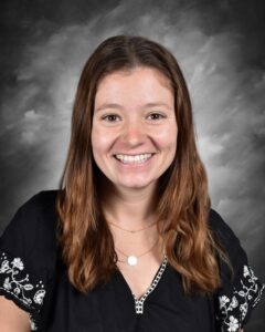 Smiling woman with long brown hair, wearing a black top with white embroidery, against a gray background.