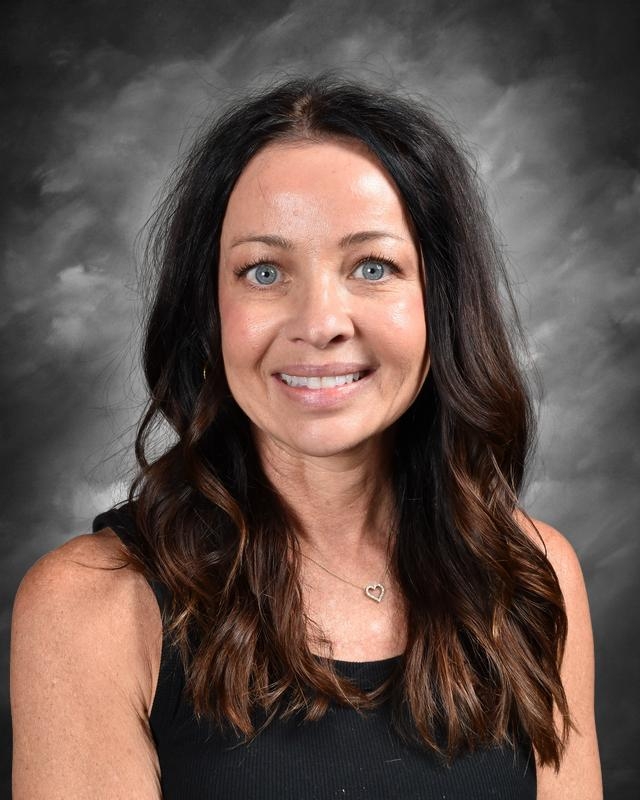 A woman with long, wavy dark hair and blue eyes smiles at the camera, wearing a black tank top and a heart necklace.