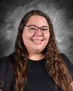 A woman with long, wavy hair and glasses smiles in front of a gray backdrop, wearing a black shirt with puffed sleeves.