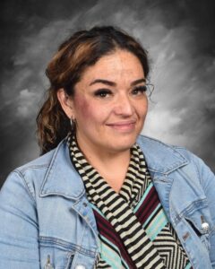 A woman with curly hair smiles, wearing a denim jacket and a striped top, against a blurred gray background.