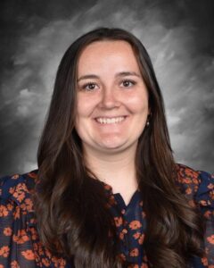 Smiling woman with long brown hair, wearing a floral blouse, against a gray background.
