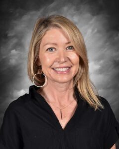 Smiling woman with long blonde hair, wearing a black shirt and hoop earrings, against a gray background.
