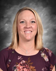 A woman with shoulder-length blonde hair smiles at the camera, wearing a floral purple top against a gray background.